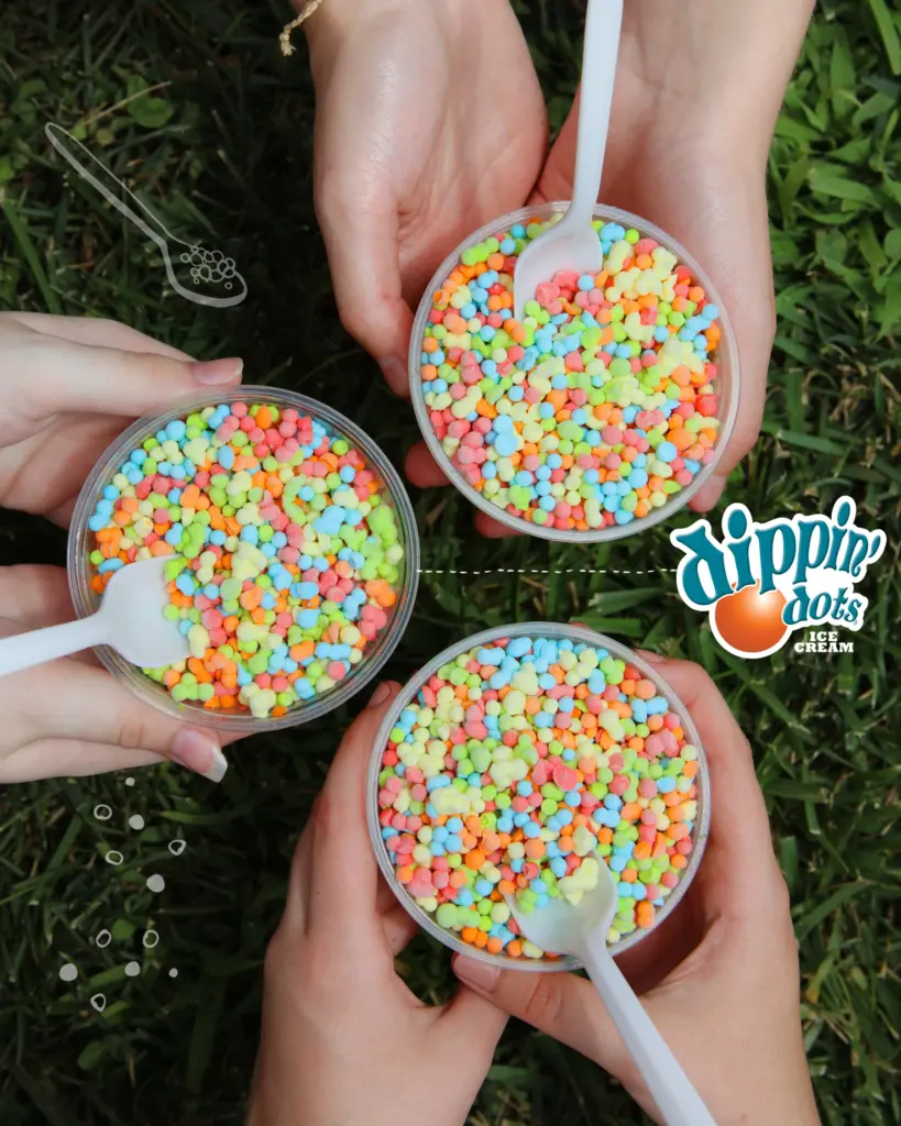 Three hands hold clear cups of colorful, beaded Dippin' Dots ice cream with white spoons over a background of green grass.