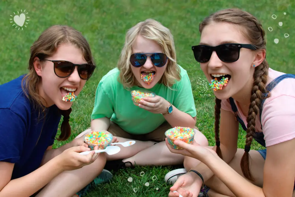 Three teenage girls wearing sunglasses sit on green grass, playfully showing off colorful Dippin' Dots on their tongues.