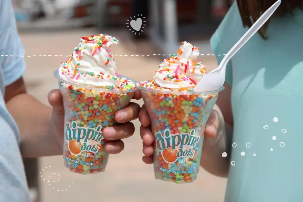 Two people hold up large cups of Dippin' Dots topped with swirls of whipped cream and colorful rainbow sprinkles.